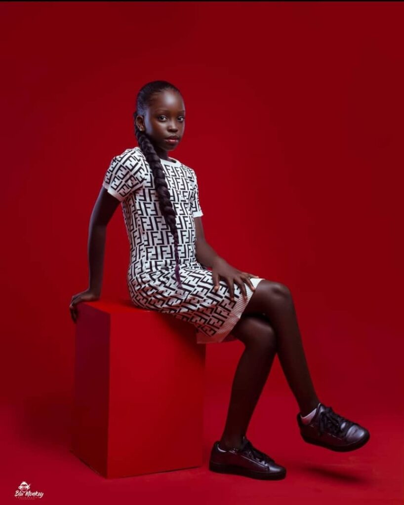 Mugasa Myra | Unravelling The Dazzling Ugandan Kid Model 5 beautiful black young girl seated on red background.