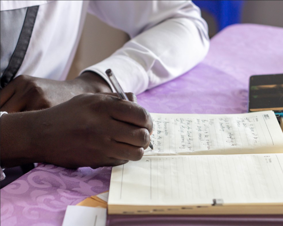 black man holds pen while writing key notes in a book and he is wearing a white long-sleeved shirt
